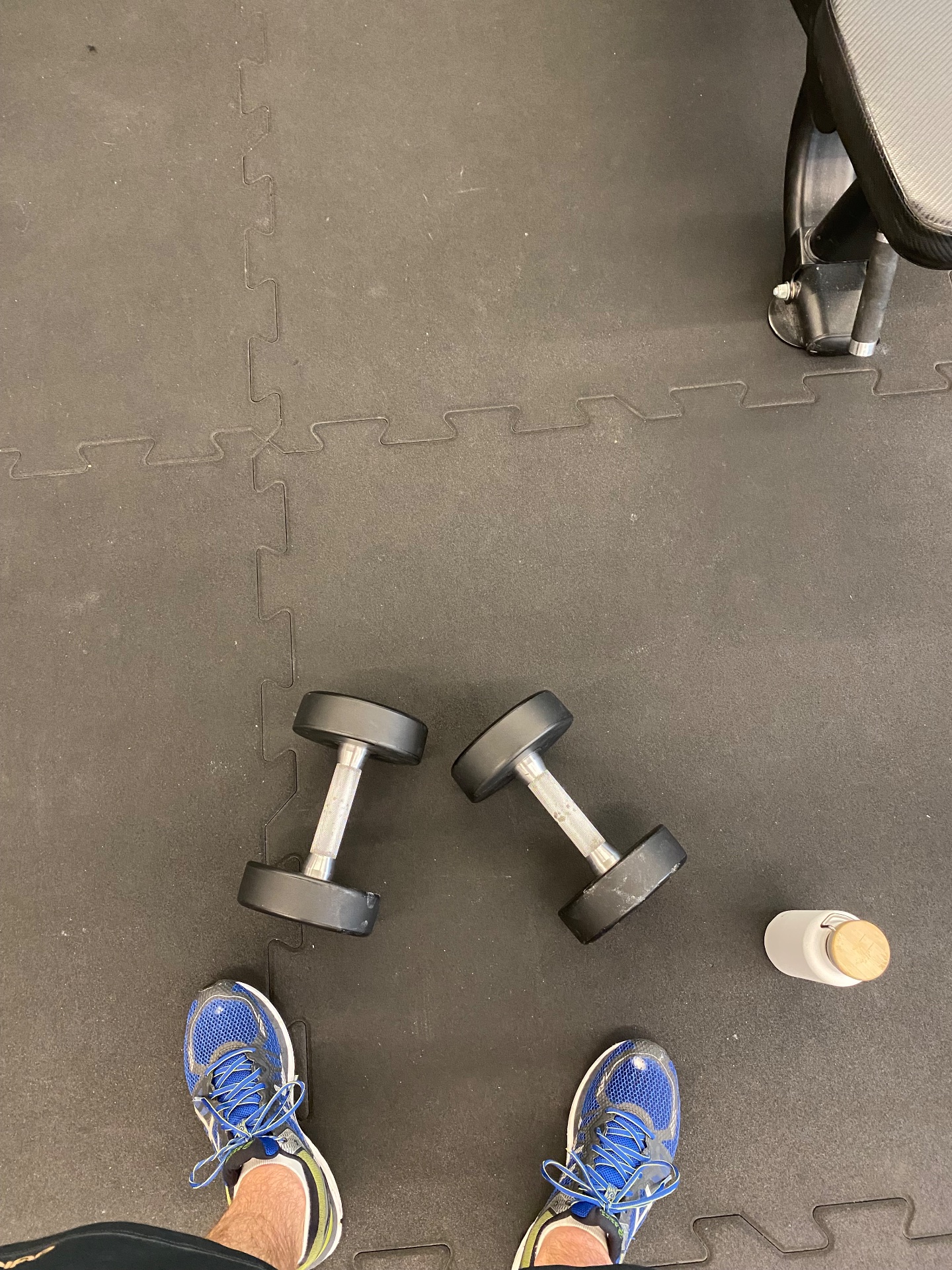 A gym floor setup showcasing dumbbells and workout gear, ready for a strength training session. The interlocking foam mat and athletic shoes suggest an intense resistance training workout is about to begin.