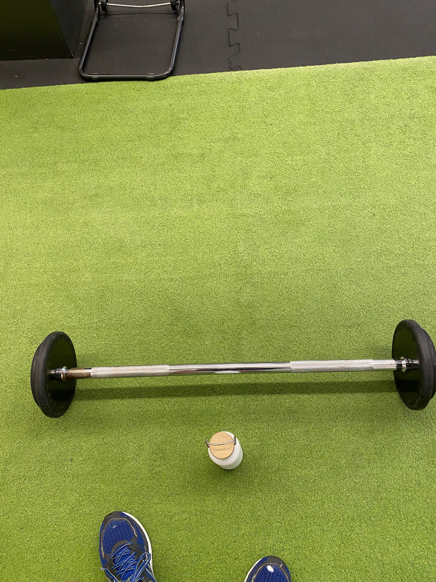 A classic strength training session with a barbell and weight plates on a gym floor. The overhead view shows someone ready to perform weightlifting exercises, with athletic shoes visible and gym equipment nearby.