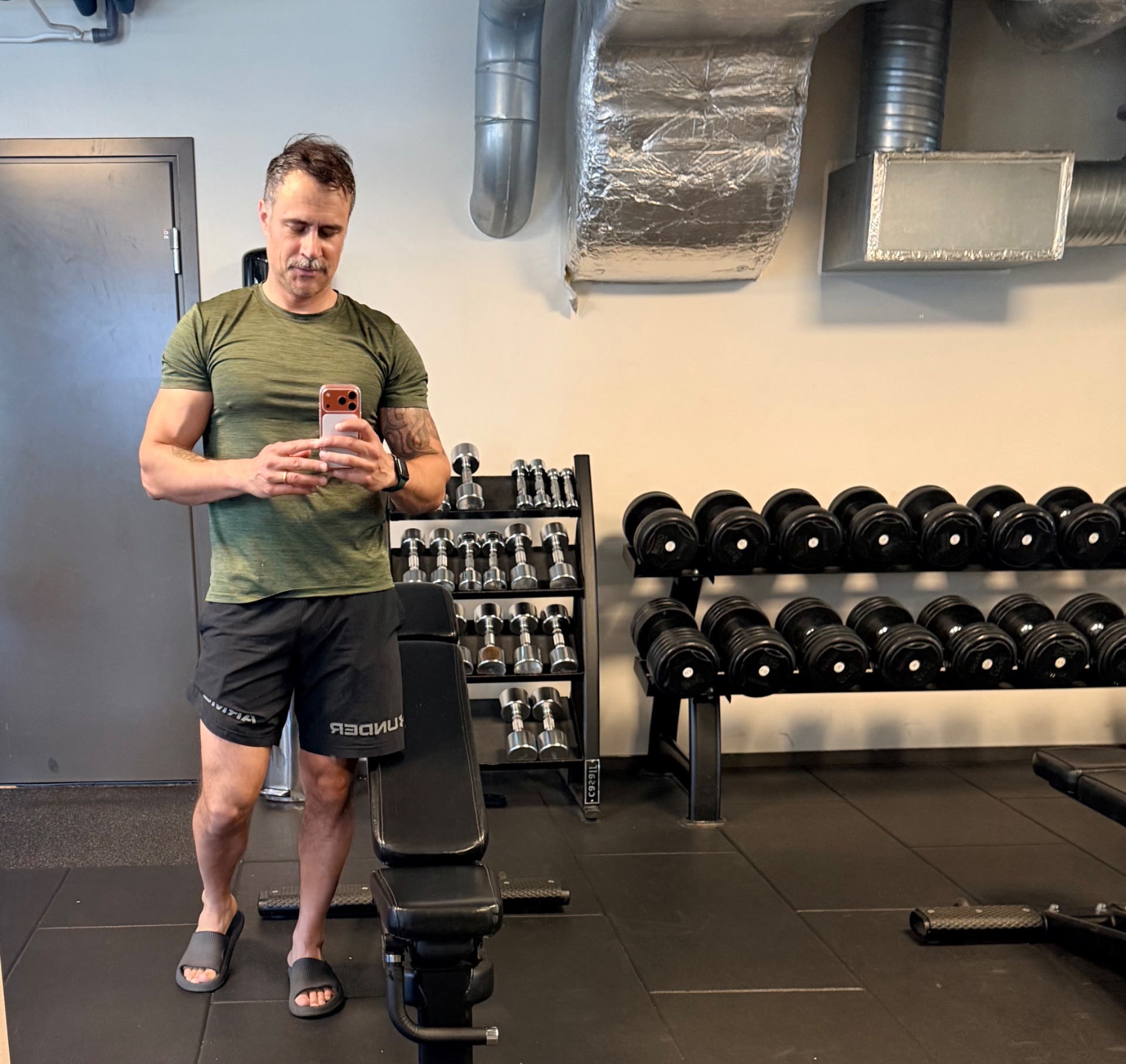 Gym selfie in a weight room with dumbbells and training equipment visible. Classic strength training setup capturing a workout moment.