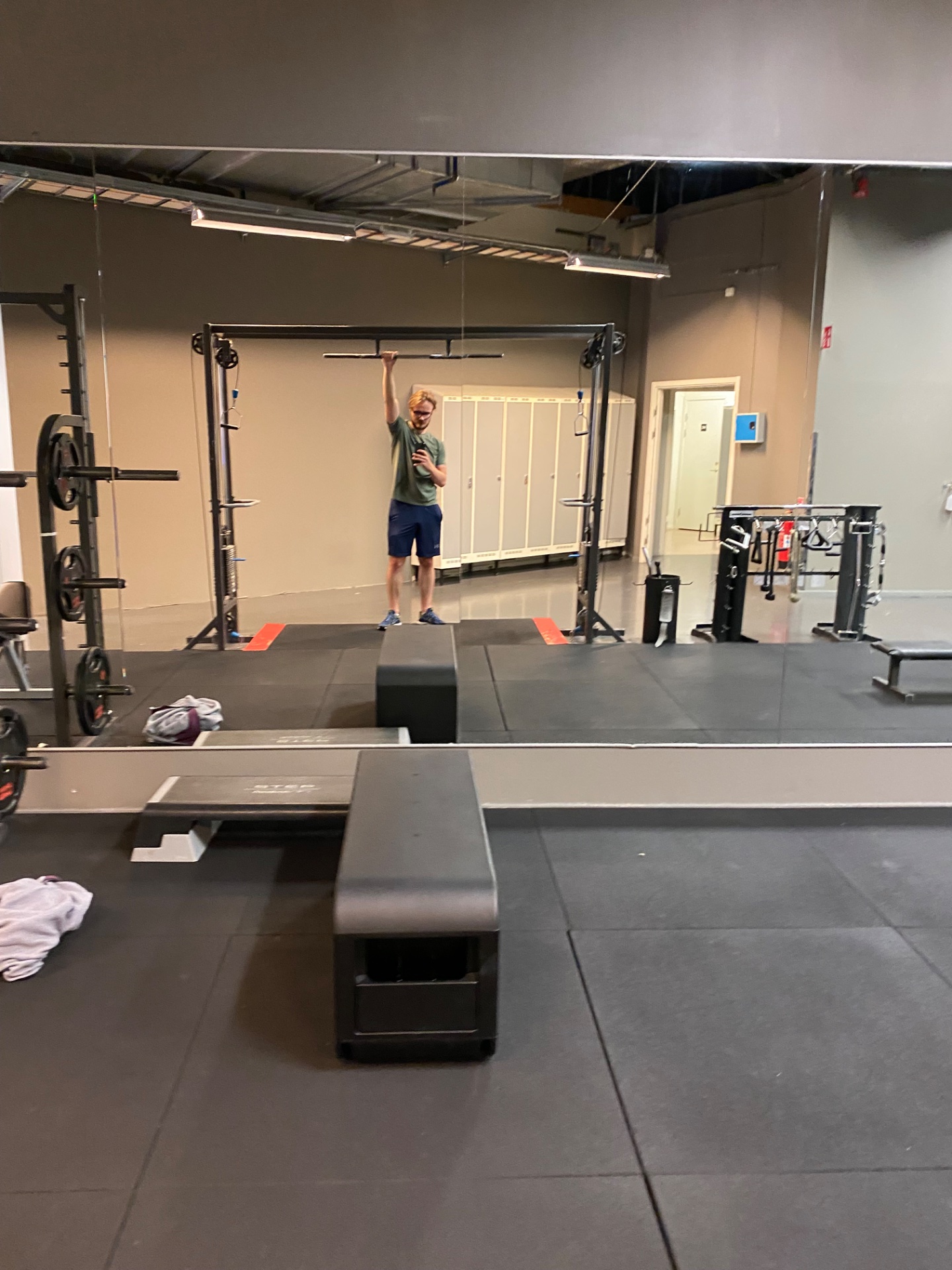A CrossFit-style gym workout featuring a person performing an overhead pull-up or lat pull exercise on a rig, surrounded by weightlifting equipment, boxes, and resistance training stations.
